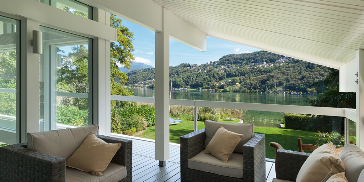 Veranda med grått tregulv og rekkverk i glass. Overbygget med hvitt tak og utsikt til grøntområde og vann.