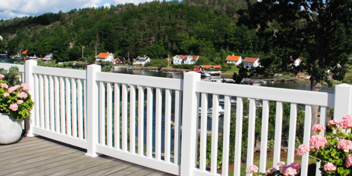Hvitt og vedlikeholdsfritt Ryvingen-gjerde montert på trebeiset verandagulv med rosa potteblomster og sjøutsikt.